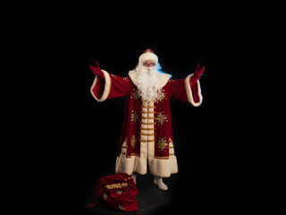 Santa Claus in a red fur coat, with a white beard and a bag of gifts posing on a black background