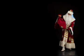 Santa Claus in a red fur coat, with a white beard and a bag of gifts posing on a black background