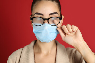 Woman wiping foggy glasses caused by wearing medical mask on red background