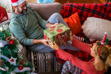 Wall Mural - Gifts share and giving action at home with happy adult couple in happy christmas holiday eve day together - red colors and love for cheerful caucasian man and woman enjoying home together holidays