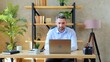 © viacheslav - Gray-haired smiling business man freelancer manager SEO sits on chair at table in home office works remote distance online uses laptop computer typing on keyboard