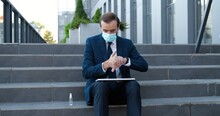 Caucasian Businessman In Suit, Tie And Medical Mask Sitting On Steps Outside, Cleaning Hands With Disinfector And Opening Laptop. Retired Man Working On Computer. Pandemic. Disinfecting. Sanitizer.