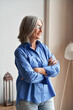 © insta_photos - Smiling confident mature woman standing arms crossed indoors looking at window. Stylish elegant middle aged senior 60s gray-haired lady wearing glasses thinking of future vision, dreaming at home.