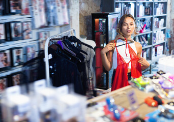 Glad cheerful positive smiling adult female purchaser touching sexy red clothes in the modern sex shop