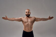Portrait of a muscular strong bald man with naked torso looking at camera while standing with outstretched arms isolated over grey background