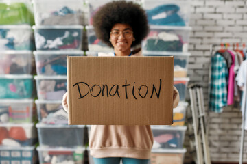 Canvas Print - African american girl holding donation box and smiling at camera, posing in front of boxes full of clothes, Young volunteer working for a charity