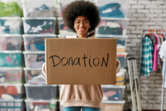 african american girl holding donation box and smiling at camera, posing in front of boxes full of c