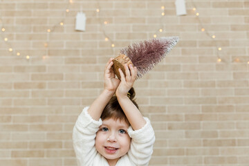 Wall Mural - Funny small girl with Christmas tree.Brick wall background.Minimalist style.Christmas light.