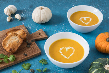 Two bowls of pumpkin soup with cream on blue background, decorated with little pumpkins, garlic and bread.