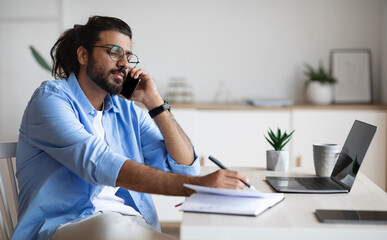 Wall Mural - Busy Western Entrepreneur Talking On Mobile Phone And Taking Notes In Office