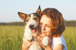 Close-up of a young woman hugs a dog in the meadow. Mixed dog with tongue smiling next to happy woman outdoors. Evening sunset outside, favorite pet.
