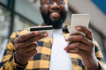 Close up of man hands holding credit card and mobile phone, shopping online. Happy African American freelancer receive payment, focus on hands