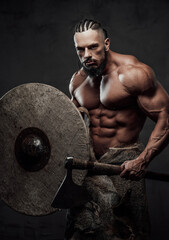 Wall Mural - Muscular guy dressed like ancient viking posing in studio with dark background holding shield and an axe with serious face.