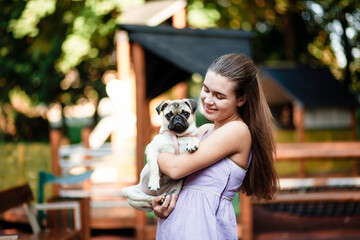 Happy dog ​​and girl. The girl is playing with a dog. Young woman walking with a pug dog in summer park. Portrait of a pug. Portrait of a beautiful pug puppy. The dog is lying on the ground.