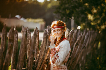 Wall Mural - A beautiful Slavic girl with long blonde hair and brown eyes in a white and red embroidered suit stands by a wooden fence.Traditional clothing of the Ukrainian region.