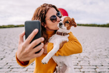 A Beautiful Woman Staying In A Park With Her Cute Little Dog. She Is Taking A Self-portrait Photo With The Phone While Kissing Her Pet. Technology Lifestyle With Pets