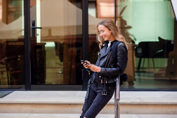 Cheerful beautiful student girl uses smart phone at stairs on the street, enjoying walk outdoors.