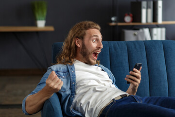 Curly-haired guy in casual style, watching great news on his mobile phone. Student receiving sms message reading good news. Surprised Man celebrating Victory on phone in Apartment.