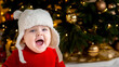 Christmas baby is smiling. A cute little girl in a red dress and white hat expresses emotions. Christmas concept with little kid, tree and garland on background in blur