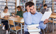 © JackF - Portrait of upset man sitting with pile of books in public library, browsing one of them