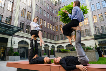 Acroyoga concept with four people, two base men lay on bench at hold at feet two flyer girls during work talking, take note, interview in office clothes.