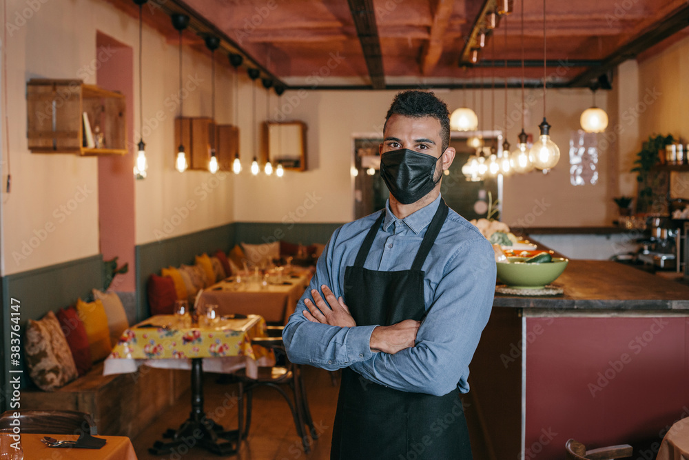 African American businessman wearing a protective black face mask and standing at his restaurant during Coronavirus. Proud owner with his arms crossed and looking at the camera with a defiant gaze. - obrazy, fototapety, plakaty 