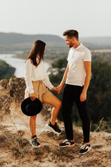 Wall Mural - Young beautiful romantic couple in stylish clothes hugging and standing on a stone on the background of a river and mountains in summer.