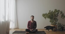 Front View Of Young Yogi Master Practicing Yoga Indoors With Cat Walking On Background. Caucasian Man Sitting In Sukhasana Pose Meditating Slow Motion. Sport Body Care Activities Healthcare 