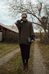 Portrait of woman in countryside. Worm clothes. Teacher. Book and eyeglasses. Autumn background. Wooden house. Village