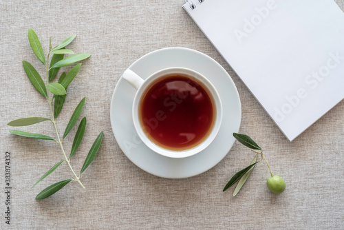 Tea break concept. Tea cup, blank notebook paper, olive branch on cotton fabric background.