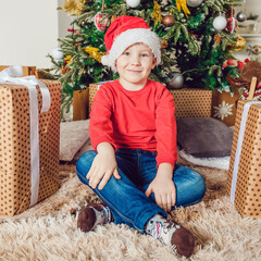 A little boy in a santa hat is sitting near a christmas tree.