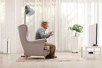 Canvas Print - Elderly man with a cup sitting in an armchair and watching tv