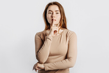 Wall Mural - Shh gesture. Young beautiful serious girl holding a finger to her lips on a white isolated background. A woman asks to be silent, a place for advertising. Negative brunette in a beige jumper.