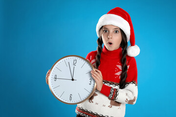 Wall Mural - Girl in Santa hat with clock on light blue background. Christmas countdown