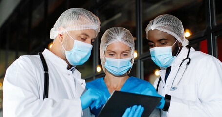 Wall Mural - Mixed-races males and female doctors in hats, medical masks and gloves standing in hospital, talking, using tablet device. Multi ethnic men and woman, medics discussing diagnosis covid-19. Coronavirus