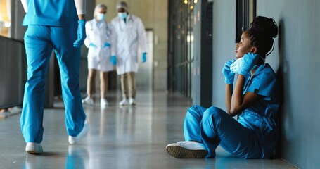 Wall Mural - Tired sad African American woman doctor in uniform and gloves taking off medical mask and leaning on wall in despair. Upset female nurse resting, having break. Coronavirus. Rest at work. Covid-19.