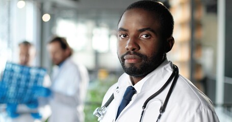 Wall Mural - Portrait of African American man physician in white gown looking at camera, smiling and standing in clinic. Handsome male doctor in hospital. Doctors on background. Medicine concept.