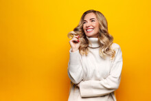 Smiling Confident Blonde Woman With Curly Hair Dressed In White Sweater, Stand With Hands Crossed, Smiles Pleasantly, Looking At Copy Space. Blank Space For Advertisement On Yellow Background. Winter 