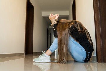 Side view unknown caucasian woman or minor girl sitting in hall of hotel with rope around her tied hands and head on knees crying desperate - Human rights kidnapping sex trafficking violence concept