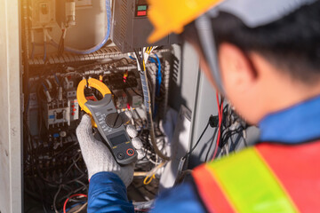 Digital clamp meter in hands of electrician close-up against background of electrical wires and relays. Adjustment of scheme of automation and control of electrical equipment.