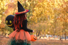 Little Girl In Witch Costume With A Balloon And A Pumpkin Bucket Playing In Autumn Park. Child Having Fun At Halloween Trick Or Treat. Kids Trick Or Treating.