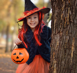 Wall Mural - Halloween trick or treat.Happy girl wearing a witch's hat, with a pumpkin handbag, peeps out from behind a tree.. Funny kid in carnival costumes outdoors.