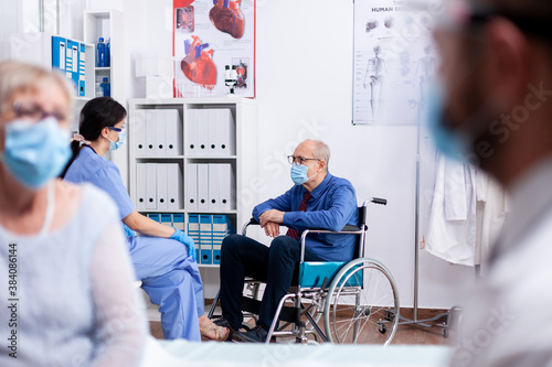 Disabled old man sitting in wheelchair wearing face mask agasint coronavirus while talking with medical staff in hospital room. Global health crisis, medical system during pandemic, sick elderly.