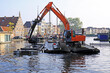A crane on a pontoon dredges a canal in the old town of|Leiden (Netherlands); dredging spoil is deposited in a transport barge.  