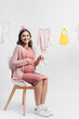 © LIGHTFIELD STUDIOS - young pregnant woman holding tiny boots and sitting near near baby clothes on clothing line on white