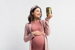 © LIGHTFIELD STUDIOS - joyful pregnant woman in headband looking at jar with pickled cucumbers isolated on white