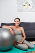 © LIGHTFIELD STUDIOS - joyful and pregnant woman touching belly while sitting near fitness ball in living room