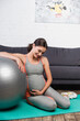 © LIGHTFIELD STUDIOS - pleased and pregnant woman touching belly while sitting near fitness ball in living room