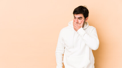 Wall Mural - Young man isolated on beige background thoughtful looking to a copy space covering mouth with hand.