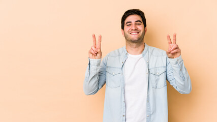 Wall Mural - Young man isolated on beige background showing victory sign and smiling broadly.